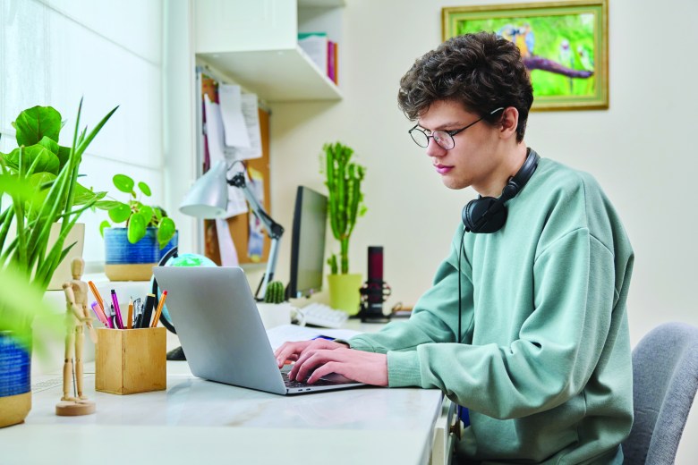 Young male college student sitting at desk at home using laptop. Handsome guy 19 years old with glasses typing on keyboard. Use internet online technologies for study, leisure, communication